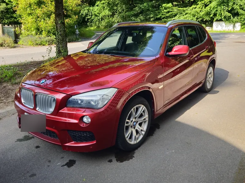 BMW X1 rouge garée au bord d'une route bordée d'arbres